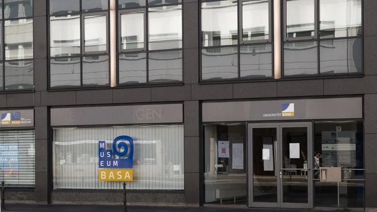 Eine Wissenschaftlerin und ein Wissenschaftler arbeiten hinter einer Glasfassade und mischen Chemikalien mit Großgeräten.