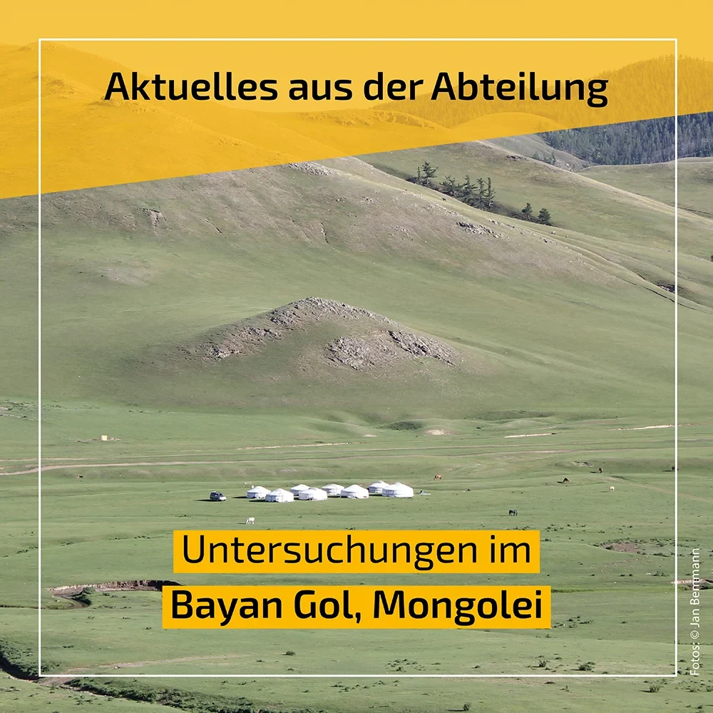 Eine Wissenschaftlerin und ein Wissenschaftler arbeiten hinter einer Glasfassade und mischen Chemikalien mit Großgeräten.